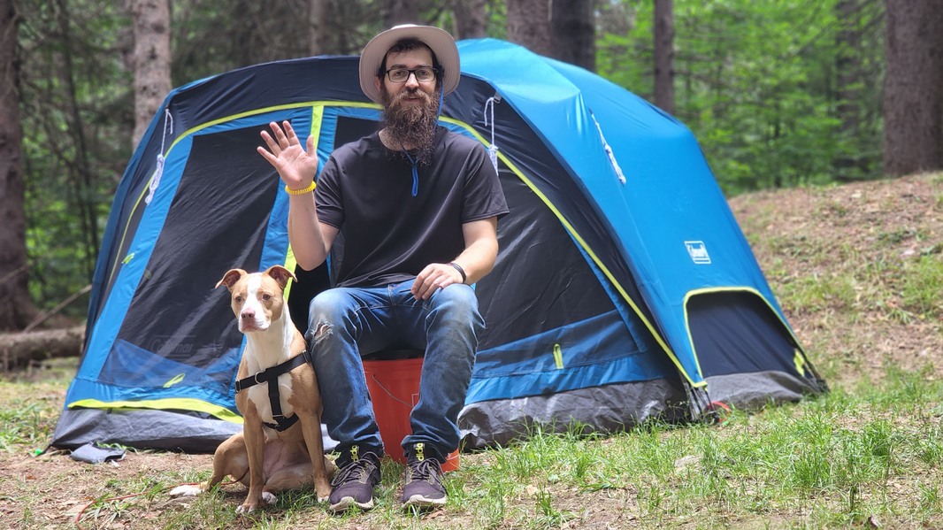 The image shows a bearded man wearing a hat sitting in front of a blue tent. The man is waving and holding the leash of his light brown and white pitbull boxer mix dog, also pictured.